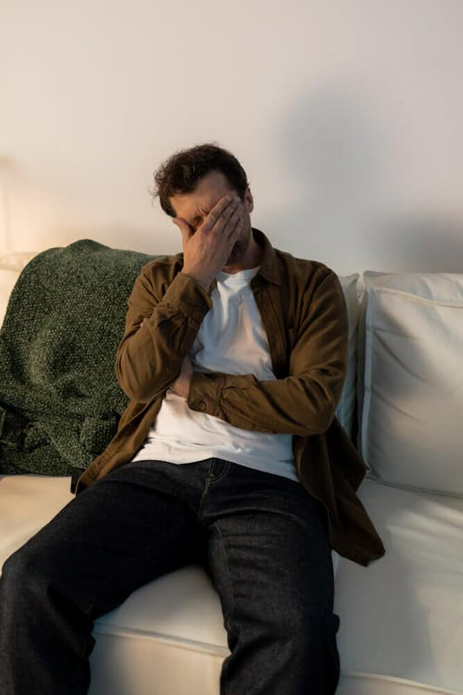 A man sitting on a couch with his head in his hand, appearing distressed.
